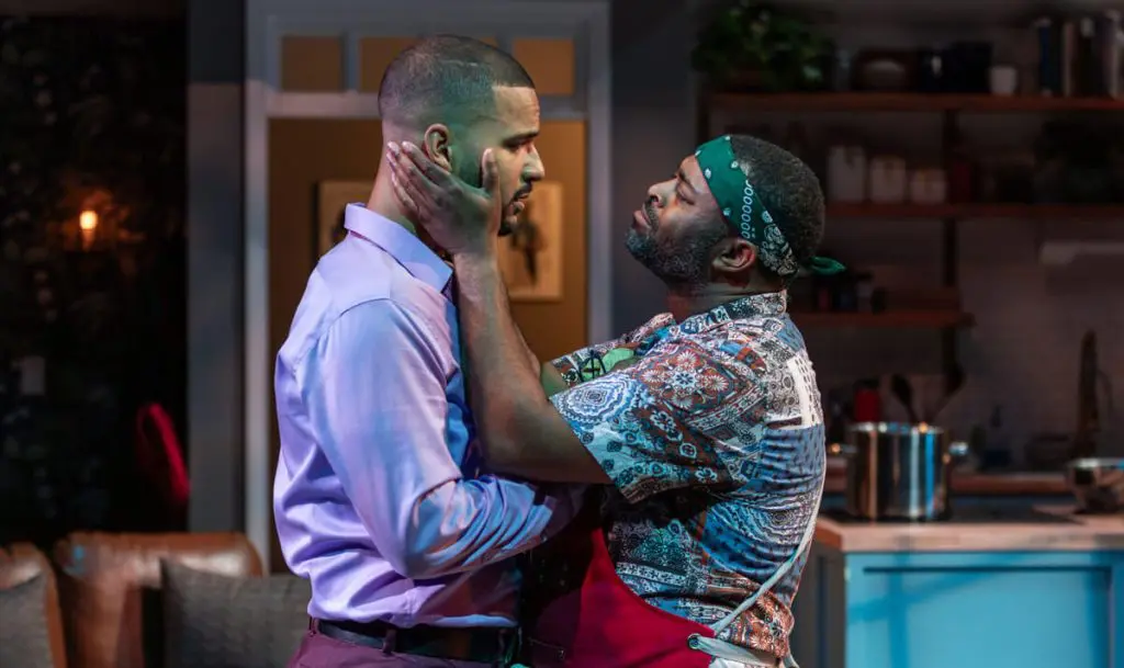 Two men stand closely facing each other in an intense, emotional moment in a kitchen setting. One man, wearing a patterned shirt and green bandana, holds the face of the other man, who is dressed in a light purple shirt and dark trousers. The scene is warmly lit, suggesting a dramatic confrontation or heartfelt exchange.