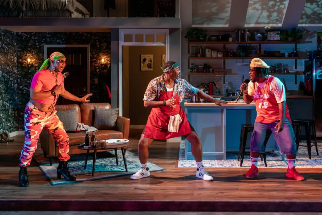 Three men perform energetically on a colorful living room and kitchen stage set. One man in a bright pink shirt and camouflage pants holds a spoon like a microphone, another man in a red apron and patterned shirt holds cooking utensils, and a third man in a red jersey and cap sings passionately into a microphone. The lively scene suggests a playful or comedic moment.