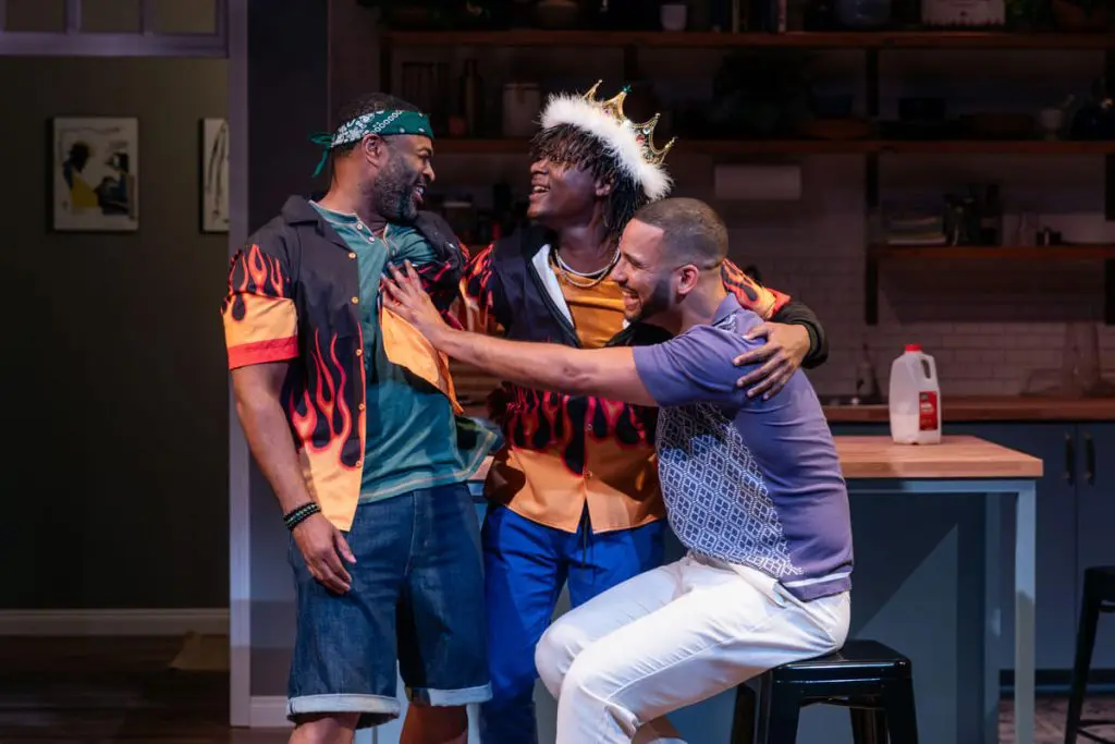 Three men share a joyful, playful moment on stage. One man in a flame-patterned shirt and denim shorts smiles warmly, while another, wearing a crown and a matching flame jacket, reaches out to him. A third man in a purple patterned shirt and white pants sits on a stool, laughing and leaning toward them. The kitchen set in the background suggests a homey, casual environment.