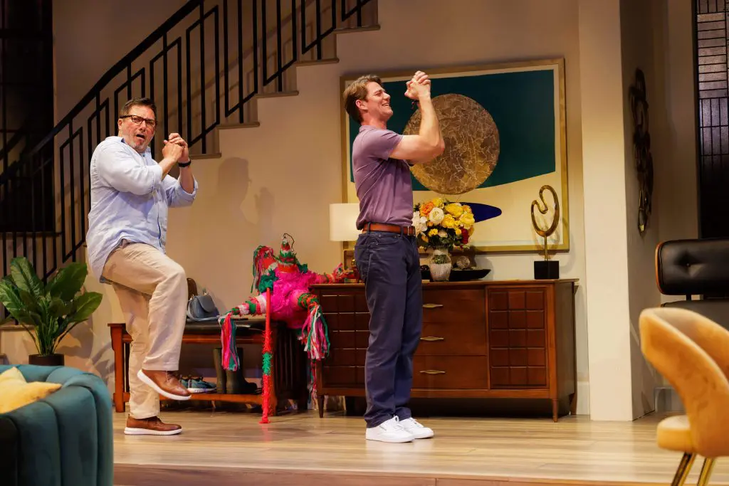 Scene from the 2025 play One of the Good Ones featuring Benito Martinez as Enrique and Nico Greetham as Marcos. In a lively moment, Enrique strikes a dramatic pose while Marcos claps enthusiastically. A colorful, broken piñata lies on the floor near a mid-century sideboard decorated with flowers and abstract art. Photo by Rich Soublet II.