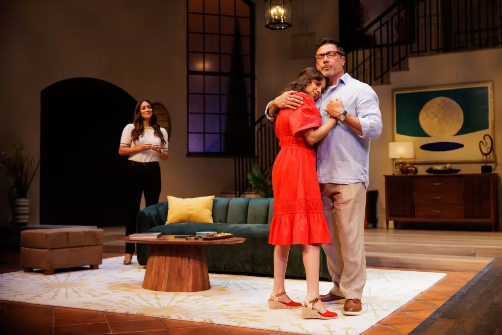 Scene from the 2025 play One of the Good Ones featuring Angelique Cabral as Ilana, Cree as Yoli, and Benito Martinez as Enrique. In a warmly lit living room set, Enrique embraces Yoli tenderly as she rests her head on his chest, while Ilana stands nearby watching them with a gentle expression. A modern staircase and contemporary art decorate the background. Photo by Rich Soublet II.