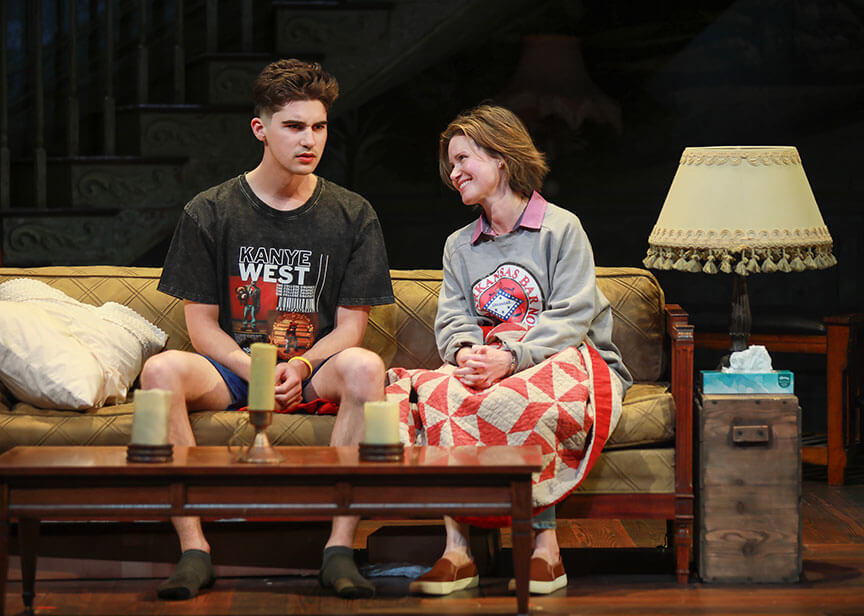 Daniel Dale and Maggie Lacey sit side by side on a worn couch in a cozy, vintage living room setting. Dale, wearing a Kanye West T-shirt and looking serious, faces Lacey, who smiles warmly while wrapped in a red-and-white quilt. A vintage lamp and wooden side table complete the intimate, emotional scene from Appropriate (2025).