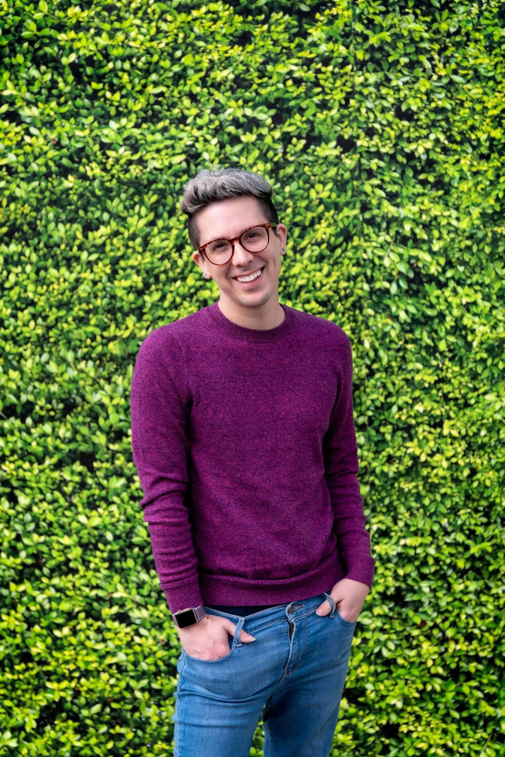 A picture of Cody Rodriguez founder of Don't Say Macbeth in a Purple Sweater and Blue Jeans with a Green Bush Background