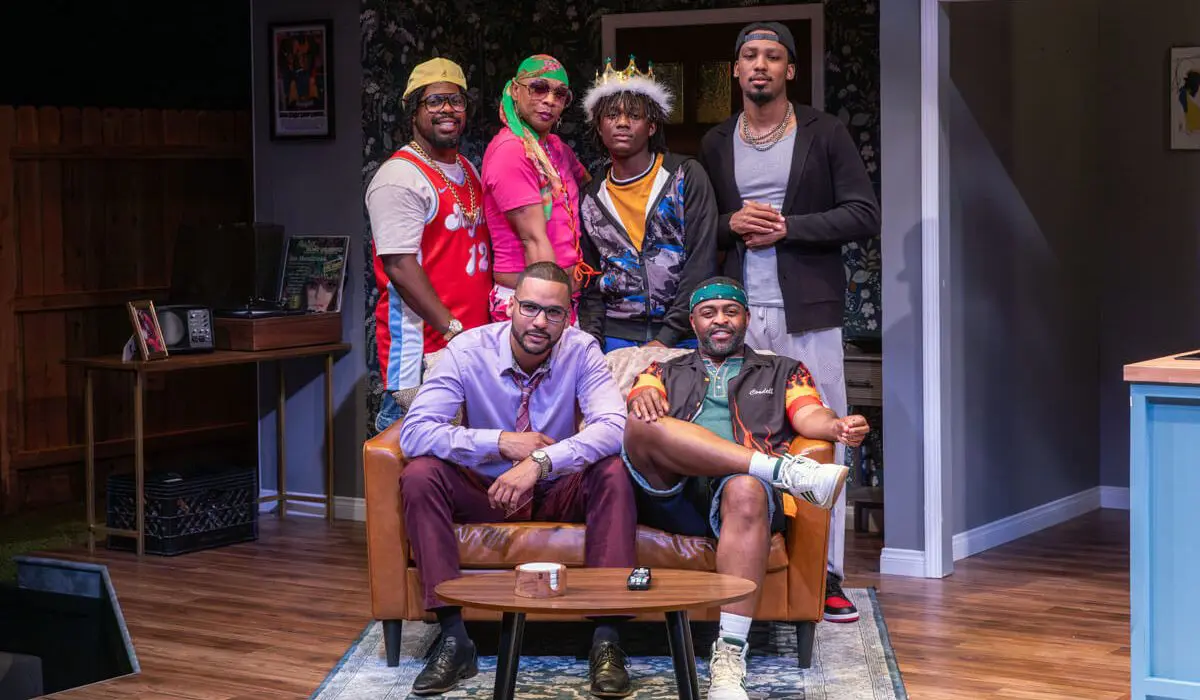 Six men pose together on a stage set designed like a cozy living room. Three men stand behind a couch, dressed in colorful and casual outfits, while two men sit on the couch — one in a light purple dress shirt and burgundy pants, the other in a sporty jacket and shorts. Another man stands confidently beside them. The setting is bright and homely, suggesting a close-knit group dynamic.