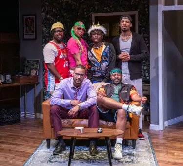 Six men pose together on a stage set designed like a cozy living room. Three men stand behind a couch, dressed in colorful and casual outfits, while two men sit on the couch — one in a light purple dress shirt and burgundy pants, the other in a sporty jacket and shorts. Another man stands confidently beside them. The setting is bright and homely, suggesting a close-knit group dynamic.