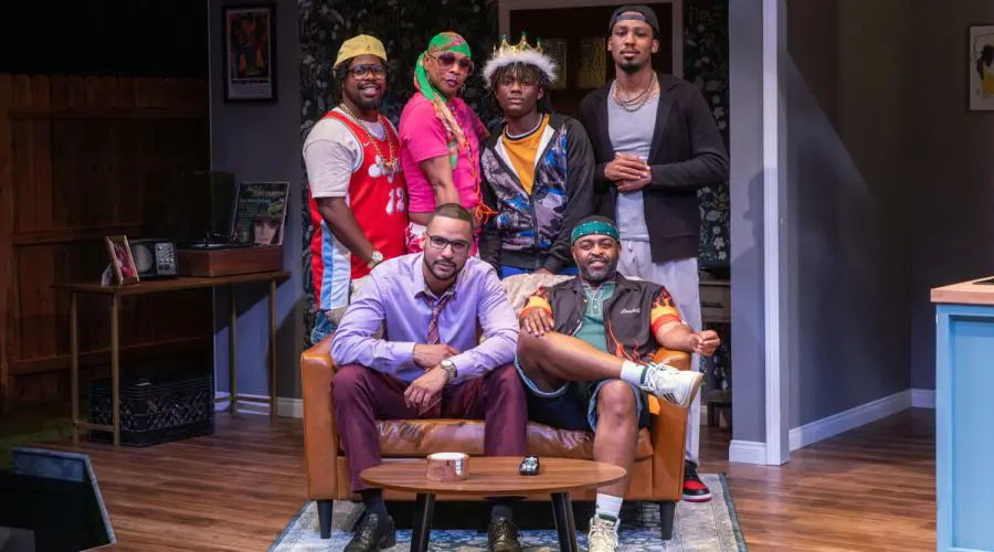 Six men pose together on a stage set designed like a cozy living room. Three men stand behind a couch, dressed in colorful and casual outfits, while two men sit on the couch — one in a light purple dress shirt and burgundy pants, the other in a sporty jacket and shorts. Another man stands confidently beside them. The setting is bright and homely, suggesting a close-knit group dynamic.