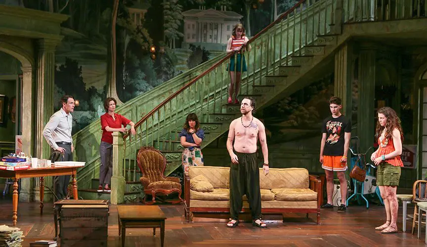 Seven actors perform on a detailed stage set resembling an old, grand Southern home with a large staircase. A shirtless man stands at the center near a worn yellow couch, surrounded by others in casual clothes. Two women lean on the staircase railing, one partway down the stairs and another higher up. Three more actors stand scattered around, watching tensely. The scene is cluttered with vintage furniture and boxes, evoking a sense of family conflict and decay.