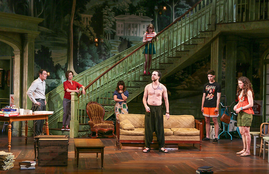 Seven actors perform on a detailed stage set resembling an old, grand Southern home with a large staircase. A shirtless man stands at the center near a worn yellow couch, surrounded by others in casual clothes. Two women lean on the staircase railing, one partway down the stairs and another higher up. Three more actors stand scattered around, watching tensely. The scene is cluttered with vintage furniture and boxes, evoking a sense of family conflict and decay.